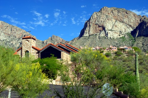 A stone church with a cross, surrounded by desert vegetation and mountains under a clear blue sky.