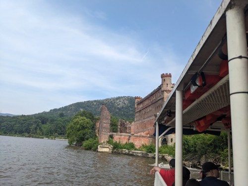 A scenic view of a castle ruins along a river, with lush green hills in the background and a boat in the foreground.