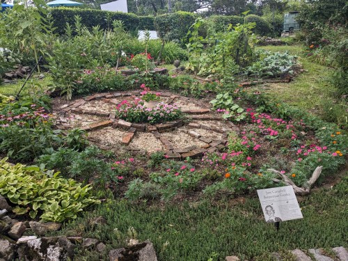 A circular garden with colorful flowers, plants, and a sign about the garden's history.