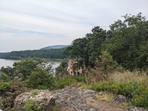 A scenic view of a river with a castle-like structure nestled among trees, surrounded by lush greenery and distant hills.