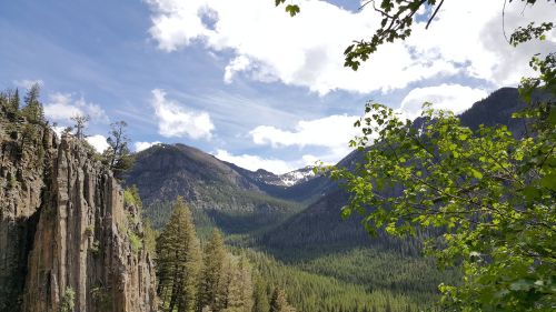 A scenic view of mountains and lush green forests under a partly cloudy sky. Snow caps visible on distant peaks.