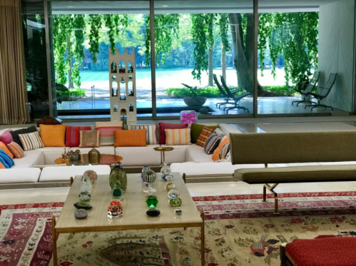 A modern living room with colorful cushions, a glass table displaying decorative items, and large windows overlooking greenery.