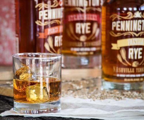 A glass of whiskey with ice, set against bottles of Pennington's Rye whiskey on a wooden surface.