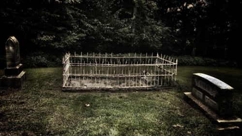 A secluded graveyard scene featuring a fenced grave surrounded by trees and two headstones nearby.