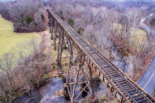 Aerial view of an old, rusty railway trestle bridge over a stream, surrounded by bare trees and grassy fields.