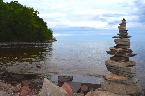A stack of stones balances on the shore of a calm lake, surrounded by trees under a cloudy sky.