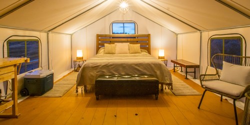 Cozy glamping tent interior with a bed, side tables, and seating, illuminated by soft lighting.