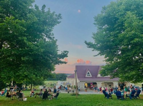 A gathering of people sitting on lawn chairs in a park, with a building and trees under a colorful sunset sky.