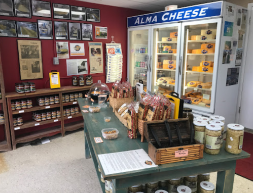 Interior of a cheese shop featuring shelves of cheese, jars, and a display case, with a red wall and photos.