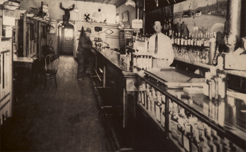A vintage bar interior with a bartender, shelves of bottles, and a mounted deer head on the wall.