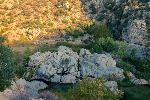 A rocky landscape with a serene pool of water surrounded by greenery and hills under warm sunlight.
