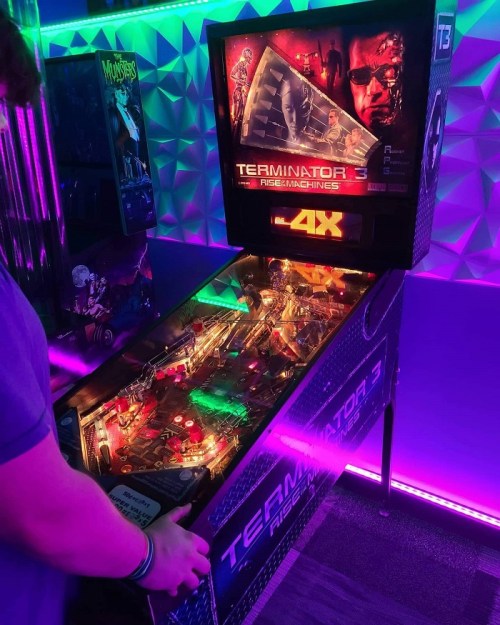 A person playing a Terminator 3 pinball machine, illuminated by colorful lights in a vibrant arcade setting.