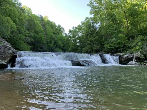 A serene waterfall cascades over rocks into a clear pool, surrounded by lush green trees and a tranquil landscape.