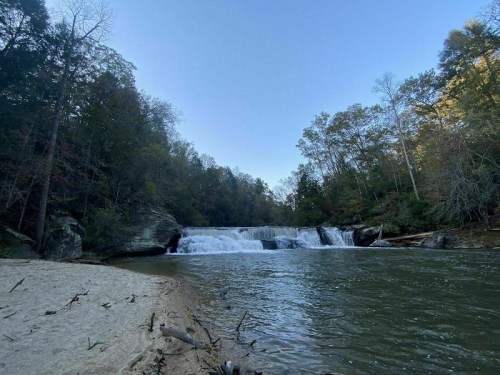 A serene river scene featuring a waterfall surrounded by trees and a sandy bank under a clear blue sky.