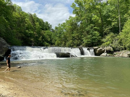 A serene waterfall cascades into a calm pool, surrounded by lush green trees and a sandy shore.
