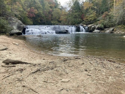 A serene waterfall cascades into a calm pool, surrounded by autumn foliage and a sandy shore.