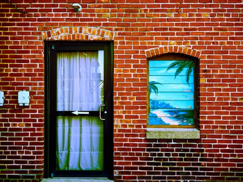 A brick wall features a door with a curtain and a window displaying a tropical landscape painting.