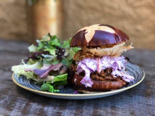 A delicious pulled pork sandwich on a pretzel bun, topped with coleslaw and crispy onions, served with a side salad.