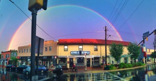 A vibrant double rainbow arcs over a colorful street scene with shops and a wet pavement after rain.