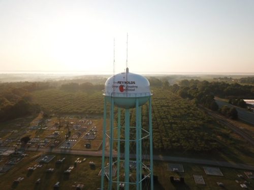 Aerial view of a water tower with "Reynolds" on it, surrounded by fields and a cemetery under a sunrise.