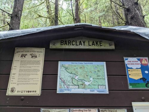 Sign for Barclay Lake with a map and bear safety information, surrounded by tall trees in a forested area.