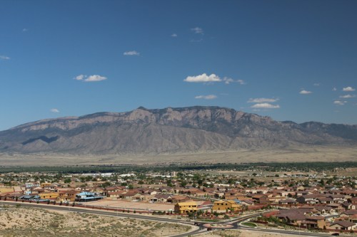 A panoramic view of a suburban area with houses in the foreground and a mountain range under a clear blue sky.