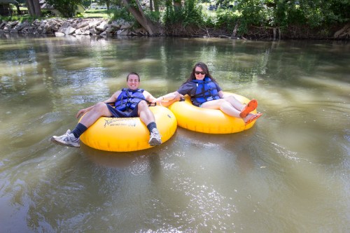 Two people relax on yellow inner tubes in a calm river, enjoying a sunny day surrounded by greenery.