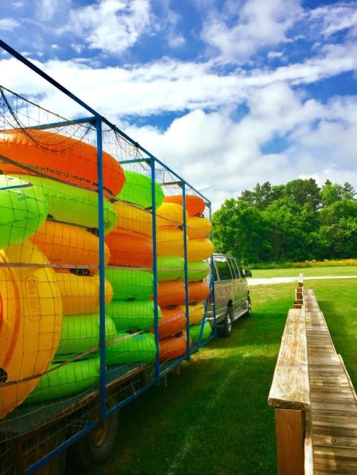 A truck loaded with colorful kayaks parked on a grassy area under a blue sky with clouds.