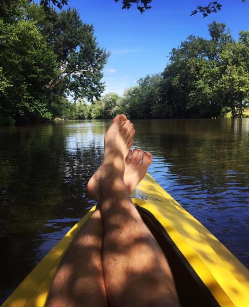 A person relaxes in a kayak, feet resting on the edge, surrounded by calm water and lush green trees.