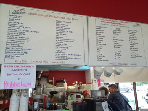 Menu board at an ice cream shop listing flavors and special recipes, with customers in the background.