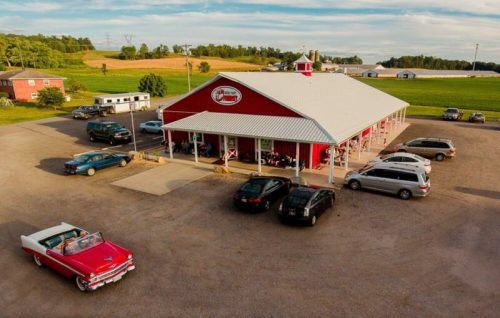 A red barn-style building with cars parked outside, set in a rural landscape with fields and a blue sky.