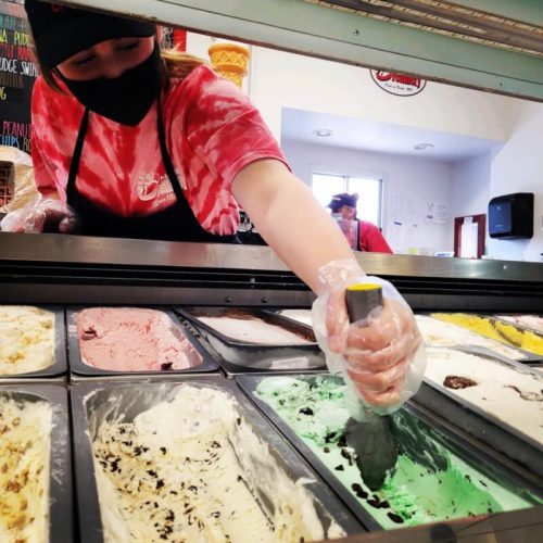 A worker in a tie-dye shirt scoops mint ice cream from a tub in an ice cream shop.