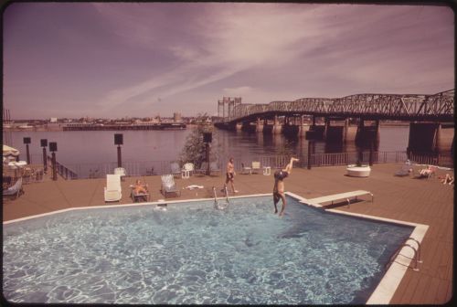 A sunny pool scene with a person diving in, lounge chairs, and a bridge over a river in the background.