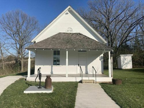 A small white building with a peaked roof, surrounded by grass and trees, featuring steps and a porch.