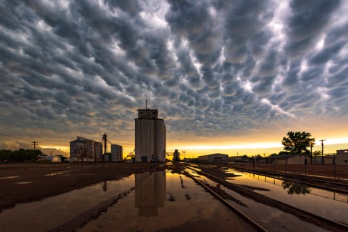 Dramatic clouds above industrial buildings reflected in puddles at sunset, creating a striking landscape scene.