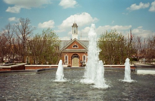 A fountain with multiple water jets in front of a brick building under a blue sky with fluffy clouds.