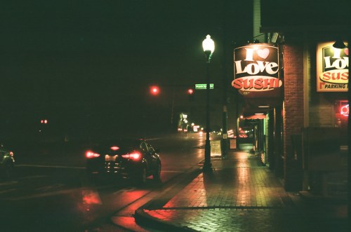 A dimly lit street at night featuring a "Love Sushi" sign and a parked car, with traffic lights in the background.
