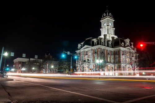 Historic clock tower building illuminated at night, with light trails from passing cars on the street.
