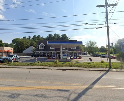A gas station with a blue canopy, surrounded by trees and parked cars, on a sunny day. Power lines in the foreground.
