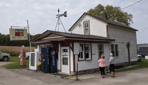 A small, vintage store with a Pepsi sign, featuring two people outside and a wind vane on the roof.