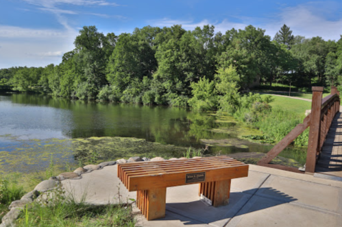 A wooden bench by a serene lake, surrounded by lush greenery and a wooden bridge in the background.