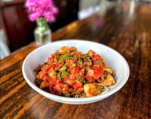 A bowl of pasta topped with a rich tomato sauce, mushrooms, and herbs, with a flower vase in the background.