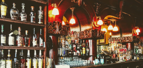A wooden bar with shelves of various liquor bottles and a well-stocked back bar, illuminated by warm lighting.