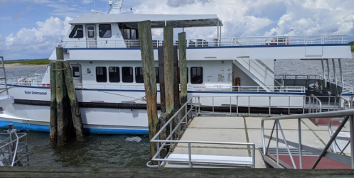 A large white ferry docked at a pier with wooden posts, surrounded by water and a cloudy sky.