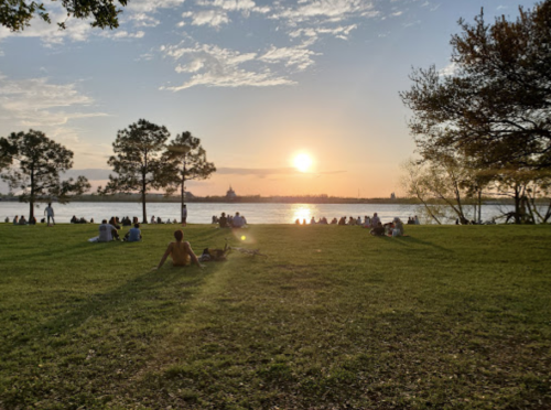 A sunset over a river, with people sitting on the grass and trees lining the shore.