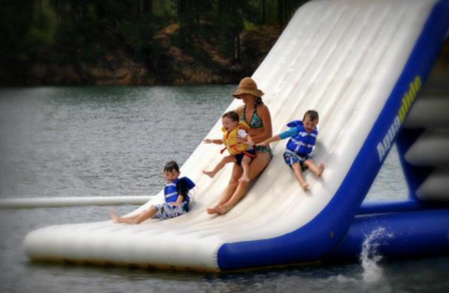 A woman and two children slide down a large inflatable water slide into a lake, enjoying a sunny day.