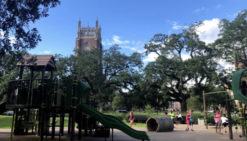 A playground with slides and climbing structures, surrounded by trees and a church tower in the background under a blue sky.