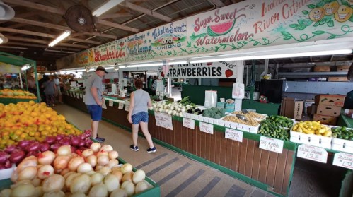 A vibrant farmers market with colorful fruits and vegetables displayed on wooden tables under a decorated ceiling.