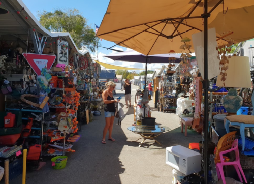 A bustling outdoor market with colorful stalls filled with various items, and shoppers browsing under large umbrellas.