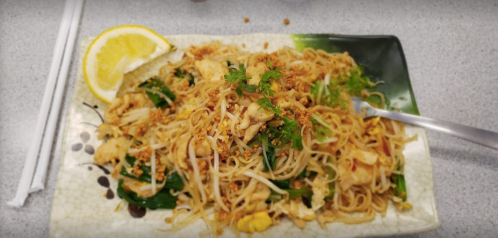 A plate of stir-fried noodles with chicken, greens, and a lemon wedge, garnished with herbs and crushed peanuts.